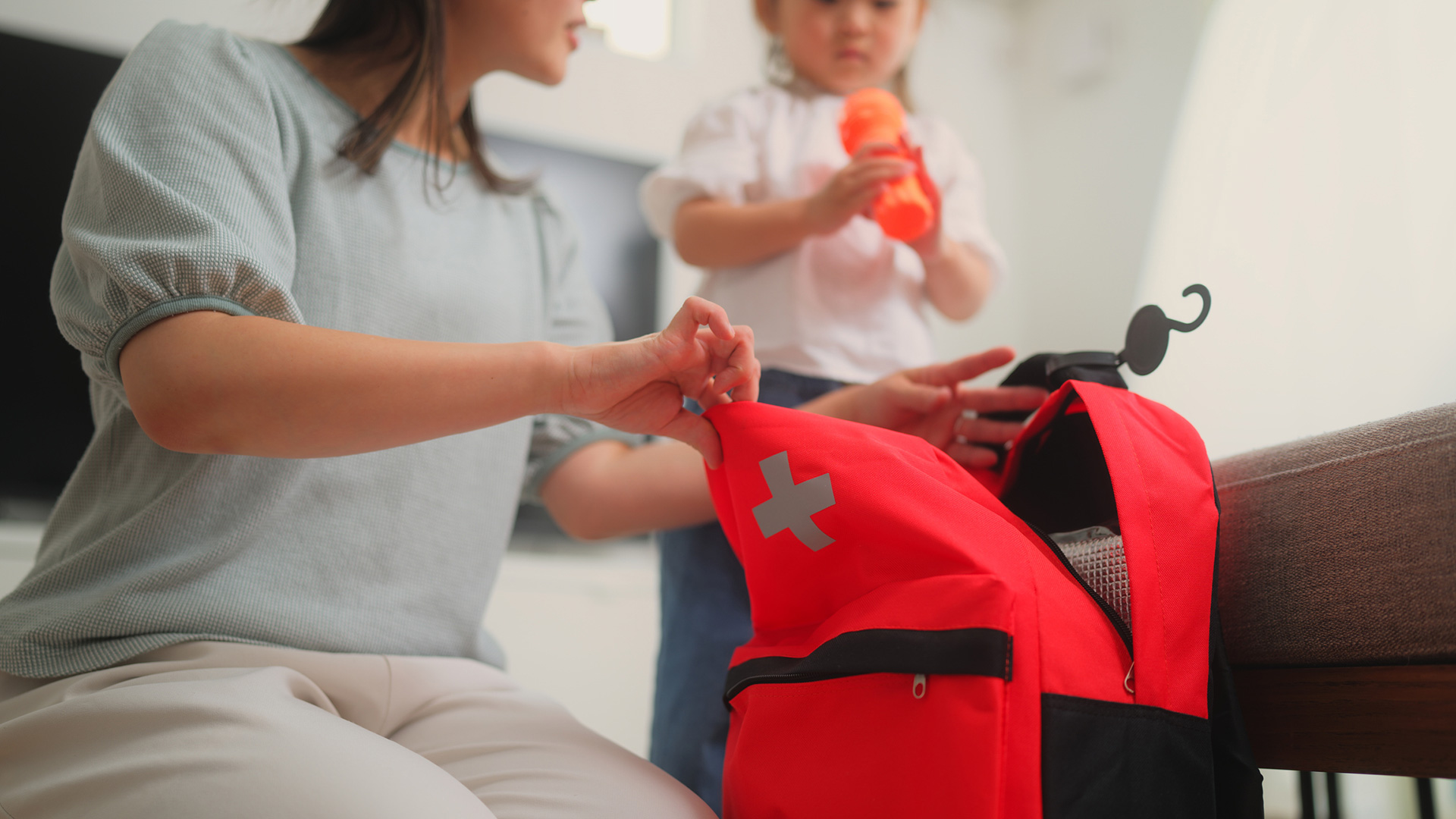 A caregiver opens a red first aid kit while a small child stands nearby holding a toy.